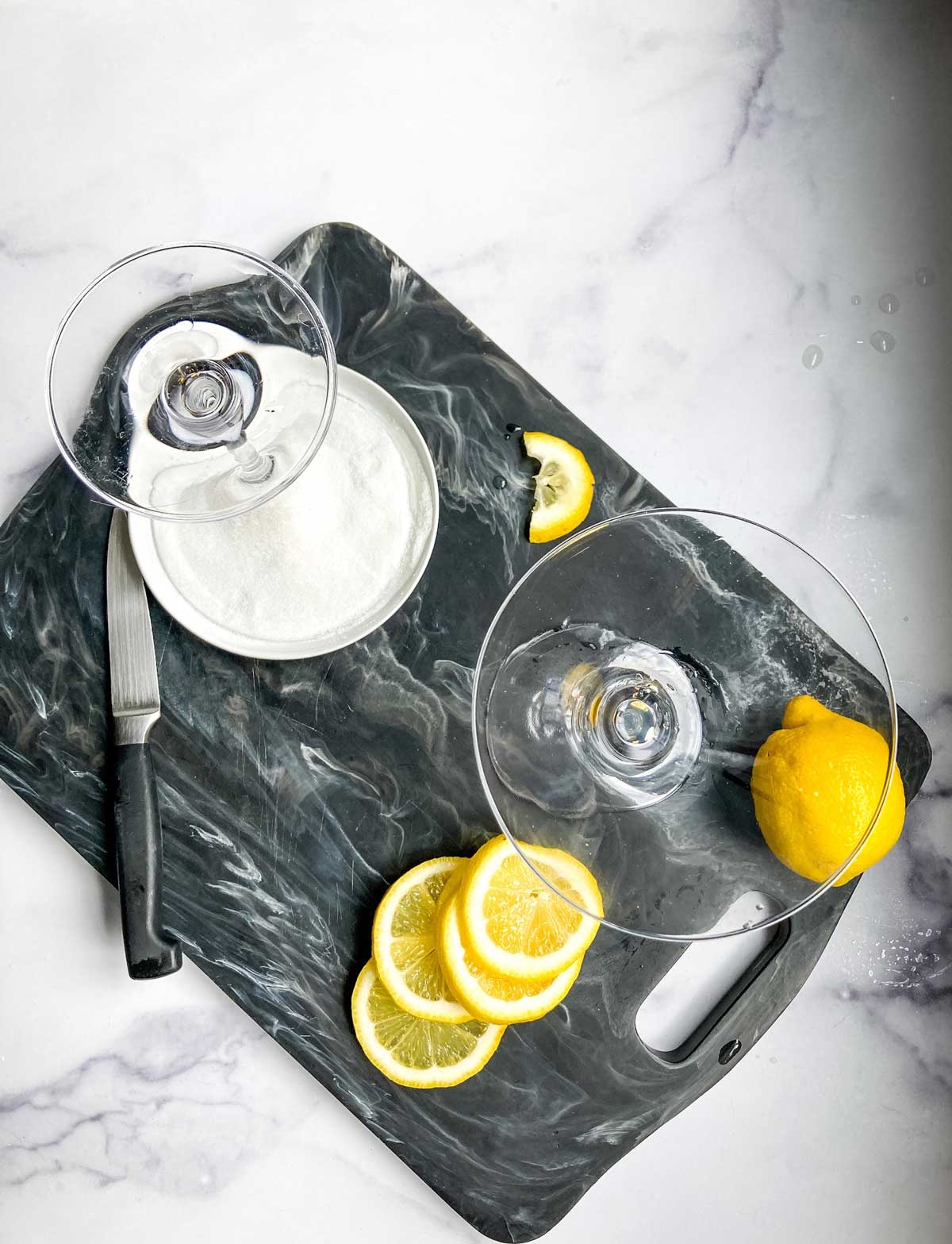 martini glasses in a saucer of sugar with lemon slices on a black marble cutting board.