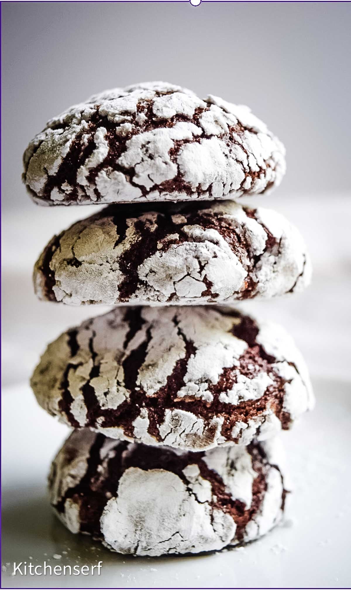 A stack of chocolate crinkle cookies coated in powdered sugar sits on a white surface.