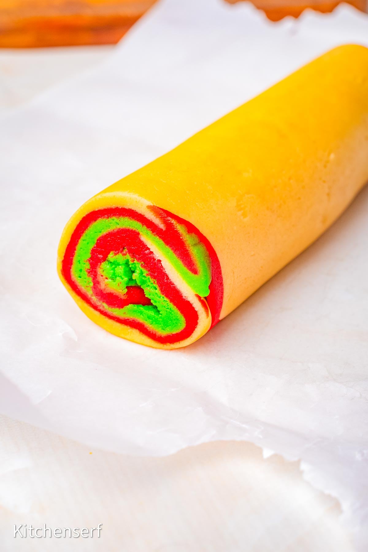 A roll of peppermint pinwheel cookie dough with red, green swirls rests on white parchment paper.