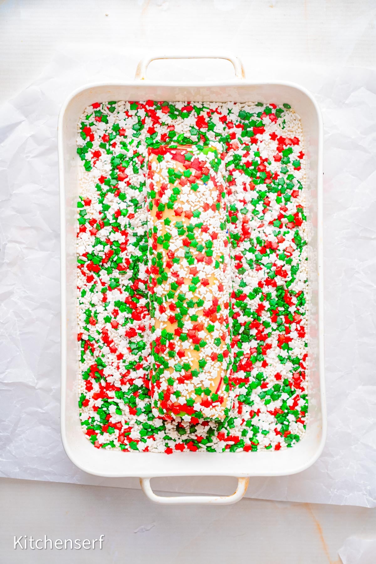 A loaf of cookie dough for peppermint pinwheel cookies, coated in red, green, and white sprinkles, sits in a white baking dish.