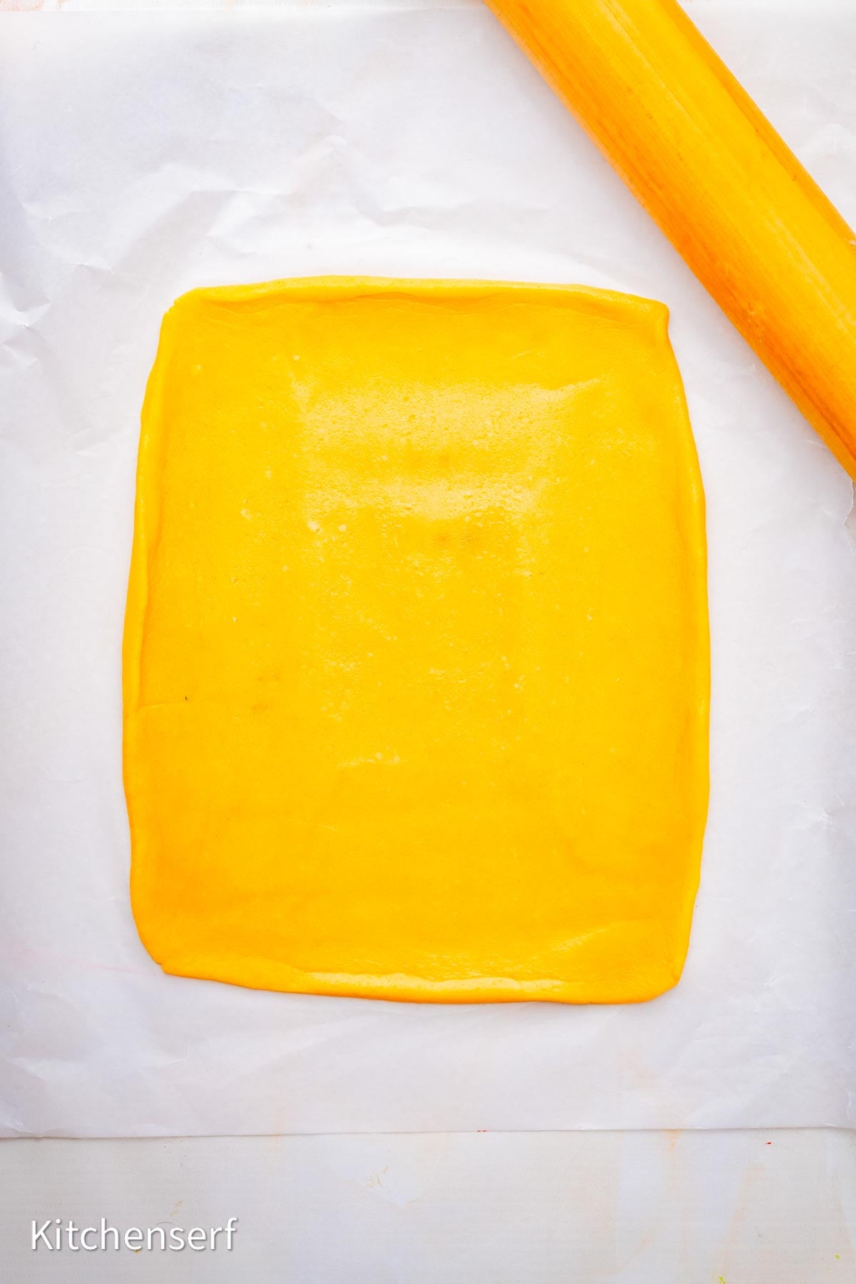 A rolled-out sugar cookie dough square on parchment paper with an orange rolling pin nearby, ready to be transformed into festive peppermint pinwheel cookies.