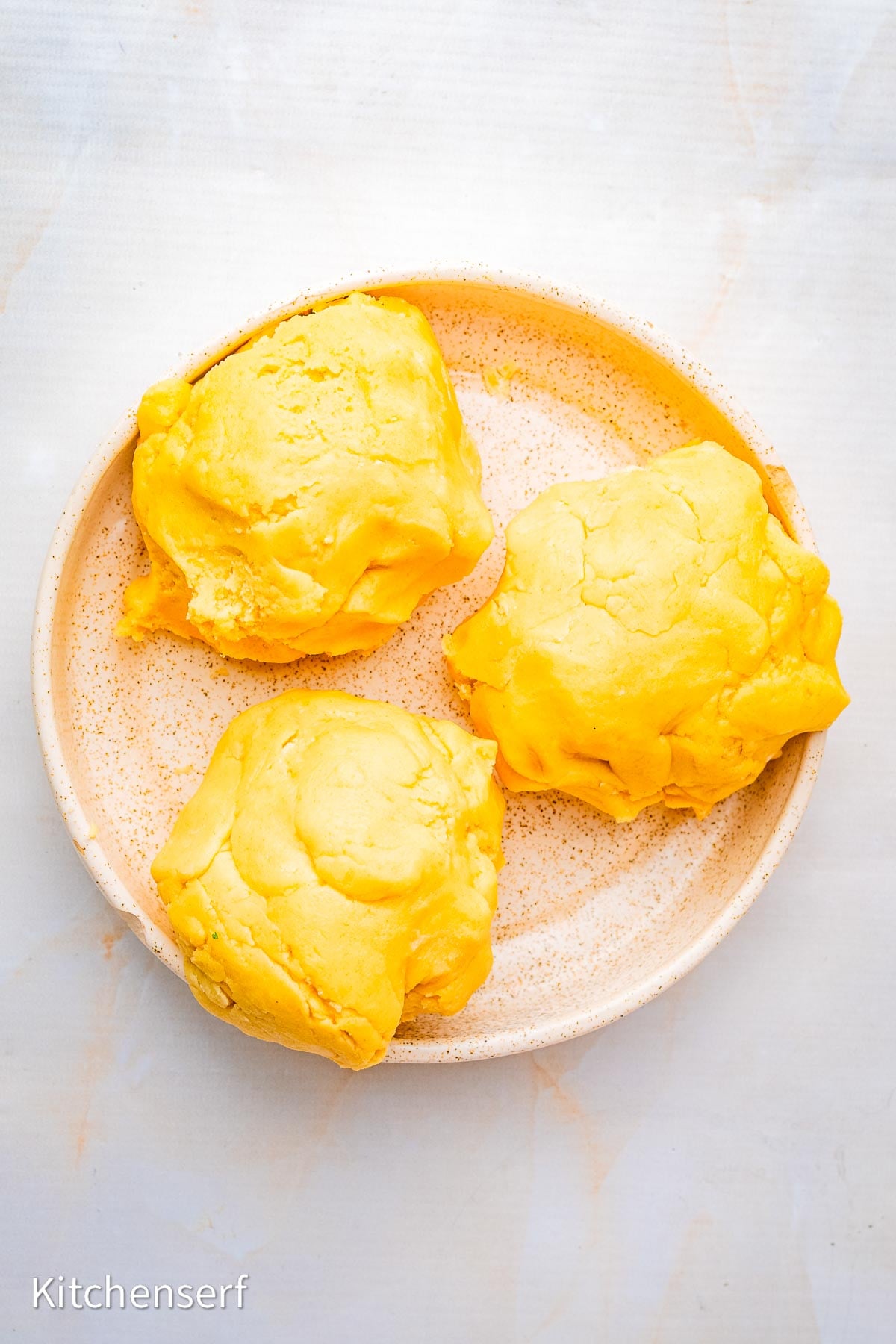 Three equal rounds of sugar cookie dough sit on a round, speckled beige plate against a light background,