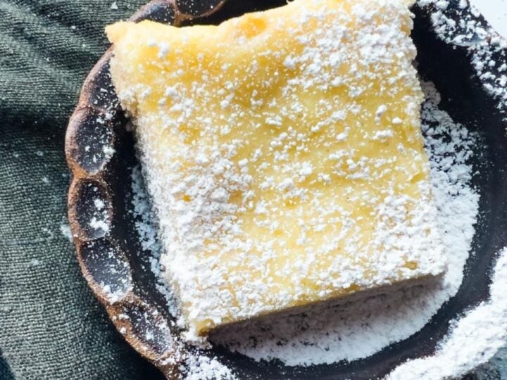 A St. Louis style gooey butter cake slice dusted with powdered sugar sits on a dark plate beside a textured green cloth.