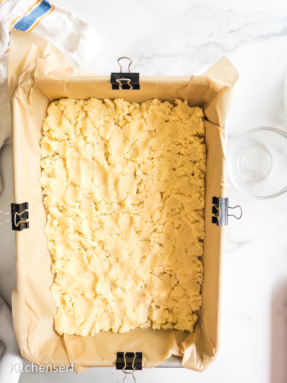 Unbaked crumbly dough, reminiscent of St. Louis gooey butter cake, is pressed into a parchment-lined rectangular pan, ready for baking.