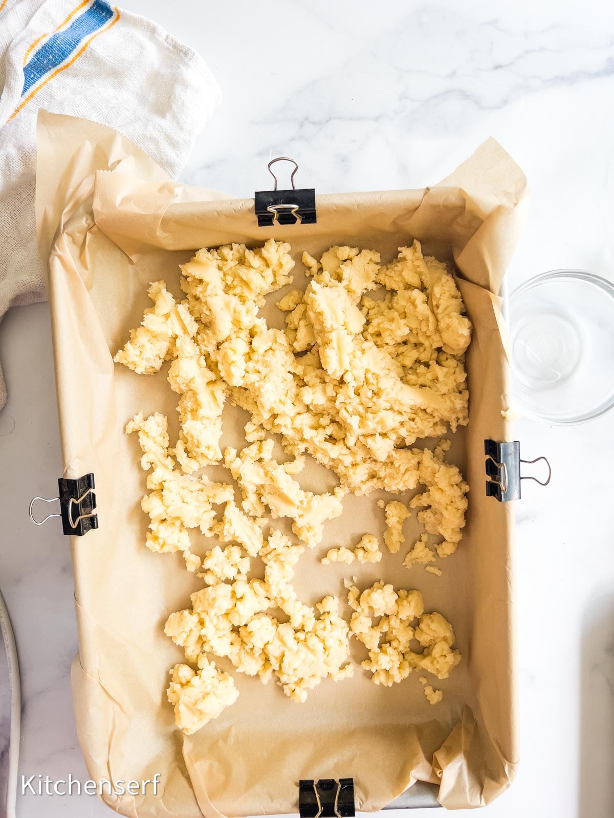 Crumbled dough for a St. Louis gooey butter cake is spread in a parchment-lined baking pan, clipped at the sides, on a white countertop.