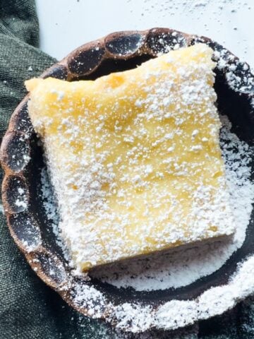 A square gooey butter cake bar dusted with powdered sugar sits on a round ceramic plate with a dark cloth beside it.