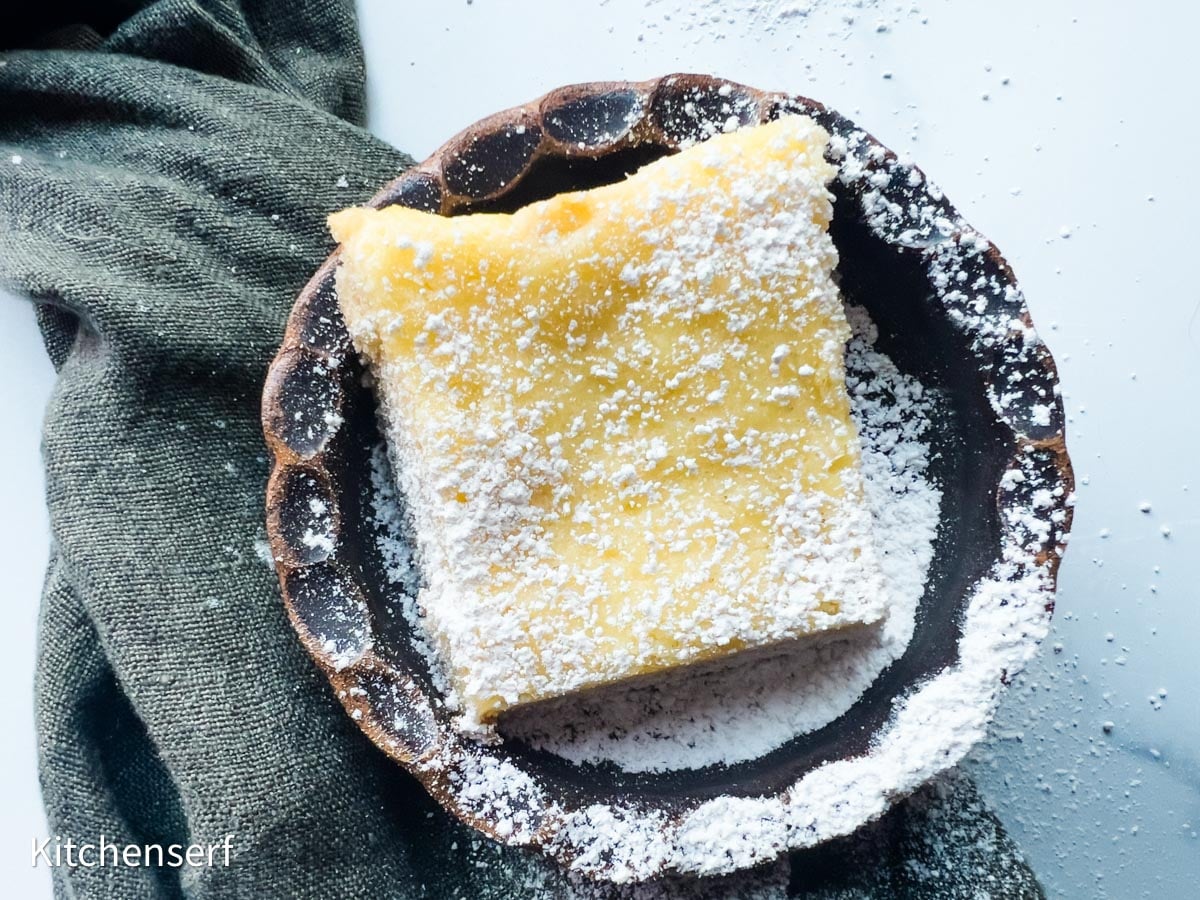 A square gooey butter cake bar dusted with powdered sugar sits on a round ceramic plate with a dark cloth beside it.