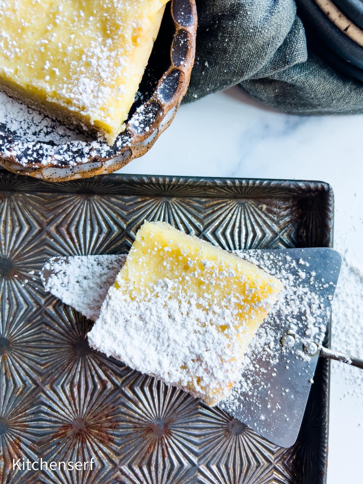 A powdered sugar-dusted slice of gooey butter cake, also known as a Neiman Marcus bar, on a metal tray.