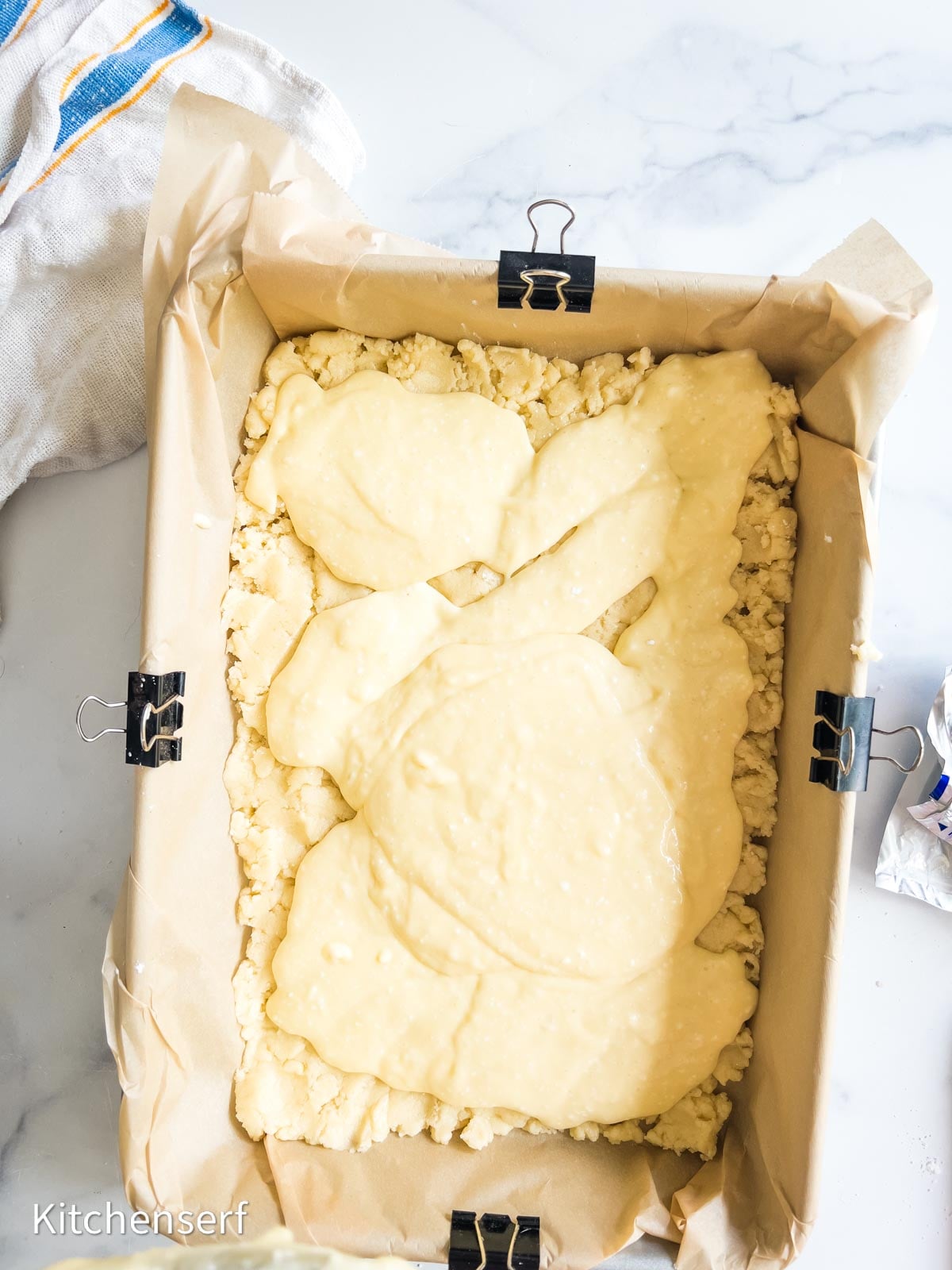 Unbaked St. Louis gooey butter cake batter spread over a crumb crust in a parchment-lined baking pan, held with binder clips.