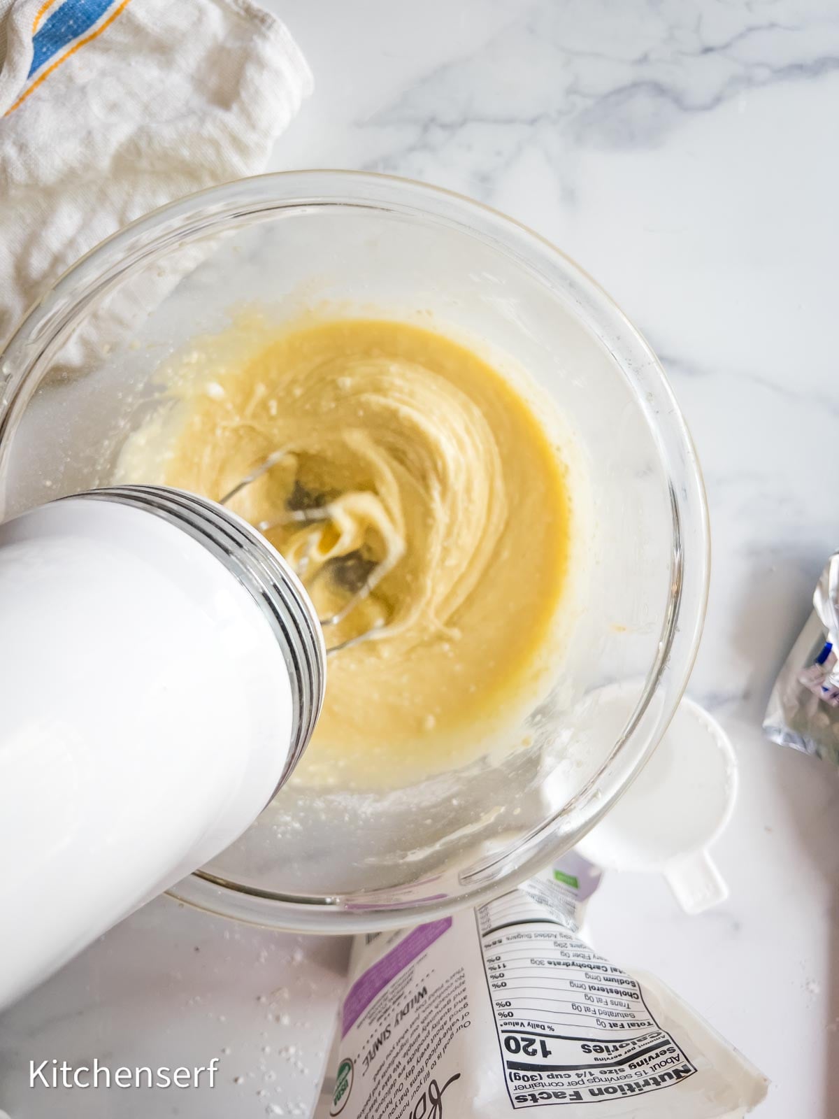 Electric mixer blending batter for a St. Louis gooey butter cake in a glass bowl on a white countertop, kitchen items around.