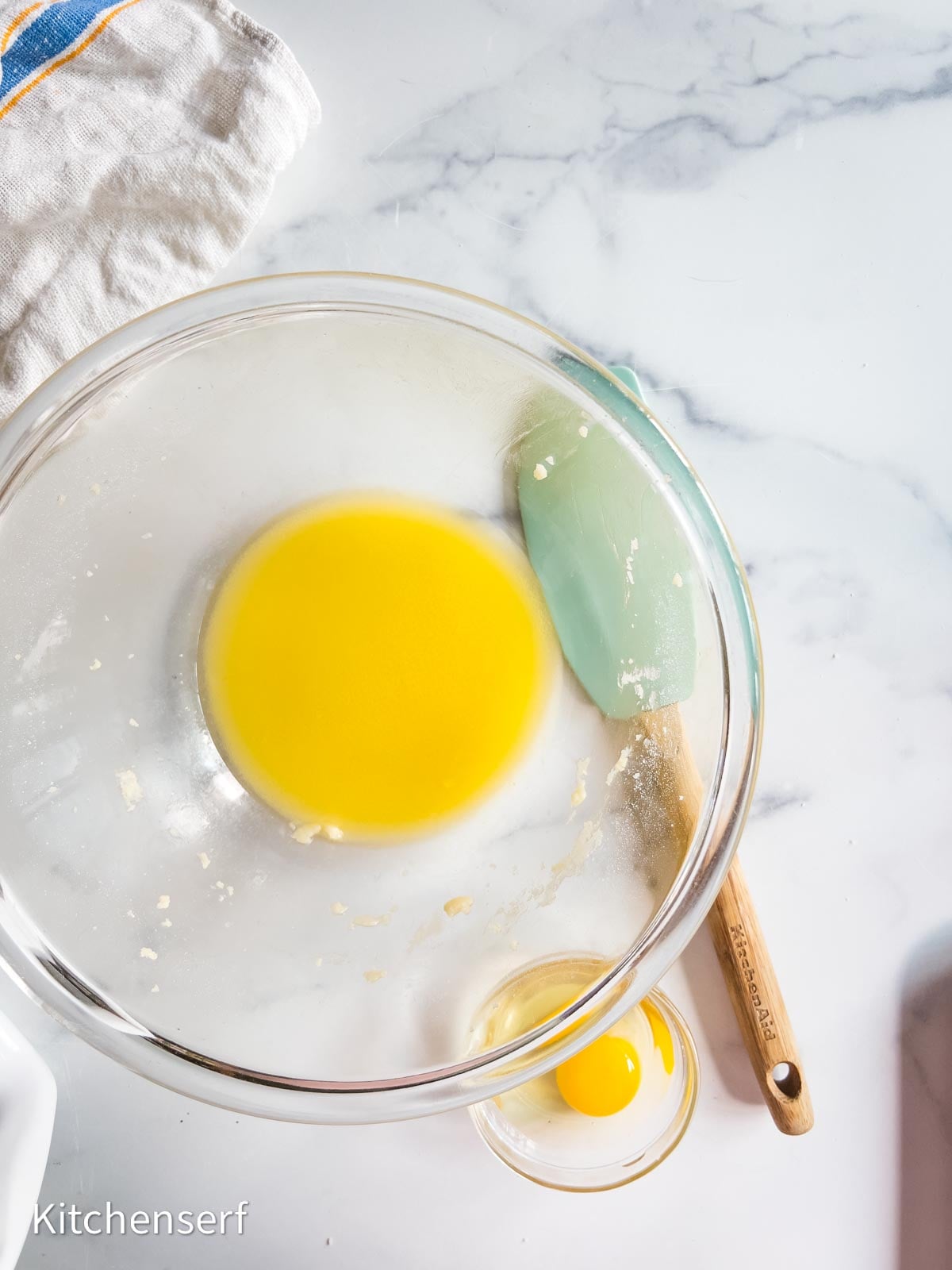 A bowl of melted butter, a silicone spatula, and a small bowl with an egg on a marble countertop—everything you need to start making a classic St. Louis gooey butter cake.