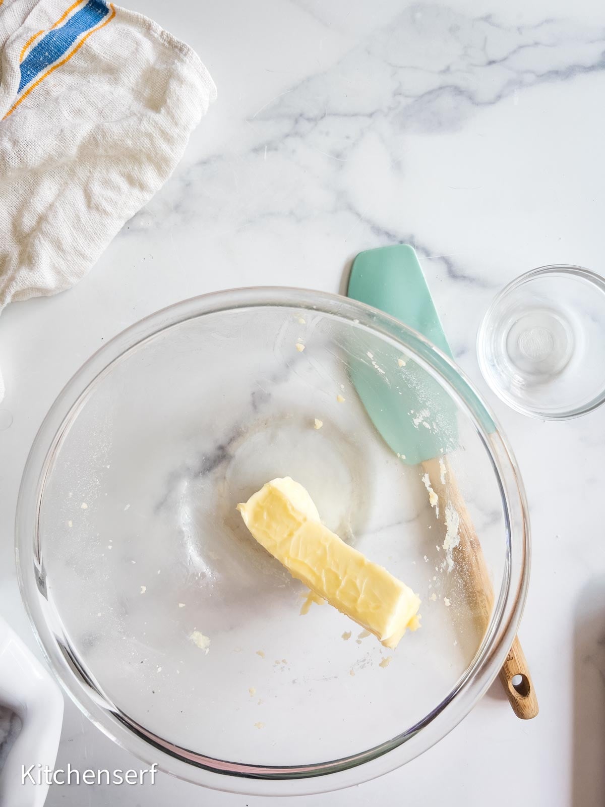 A glass bowl with a stick of butter, a spatula, a small glass bowl, and a towel on a marble counter—perfectly arranged for preparing classic St. Louis gooey butter cake.