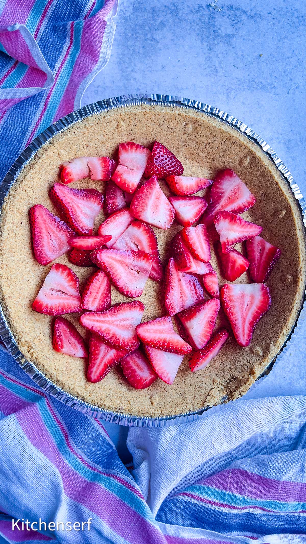 Sliced strawberries arranged on a graham cracker pie crust create a tempting strawberry cheesecake, with a striped cloth nearby.