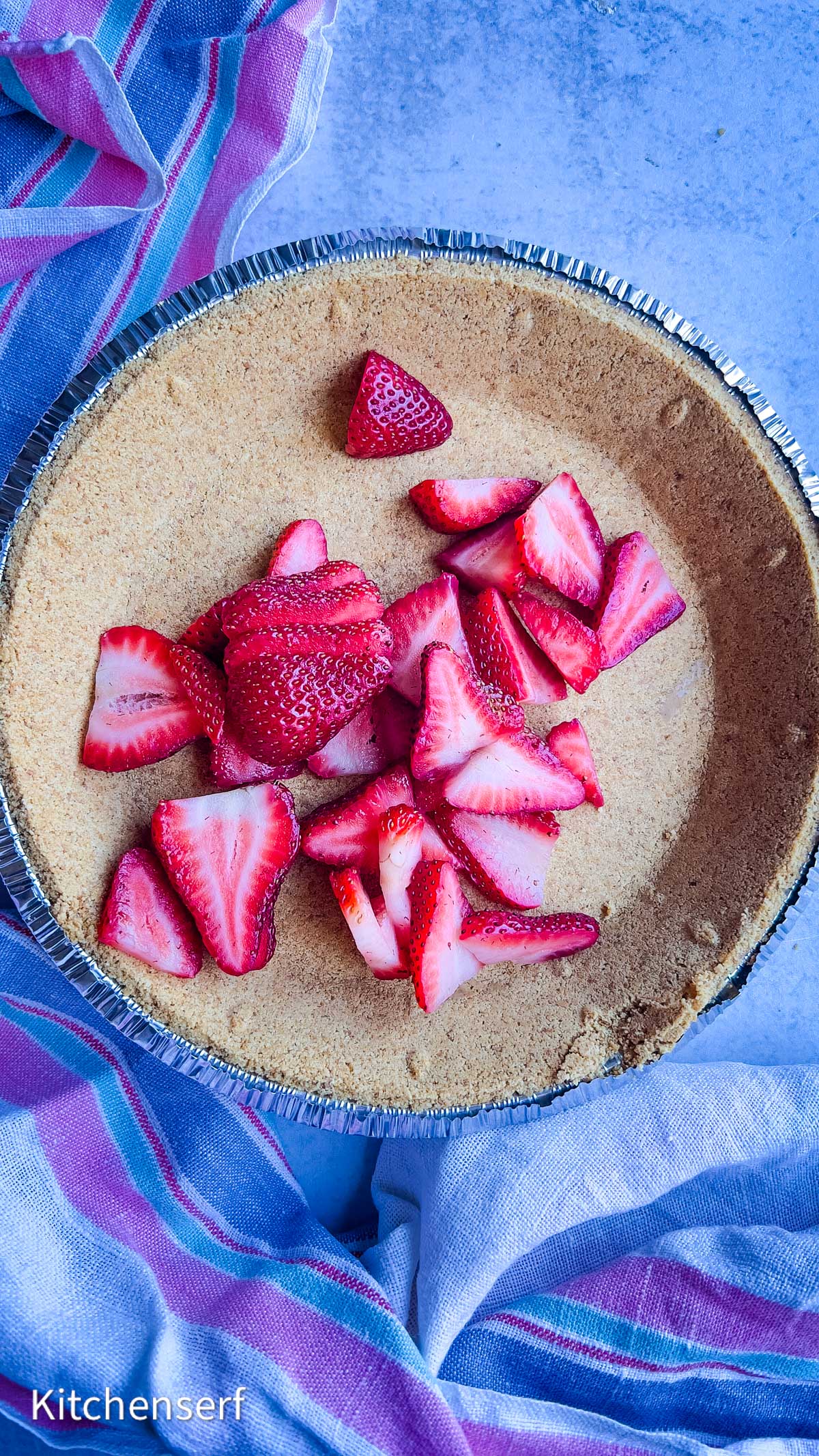Sliced strawberries in a graham cracker pie crust at a classic strawberry cheesecake, set on a blue surface with striped towels.