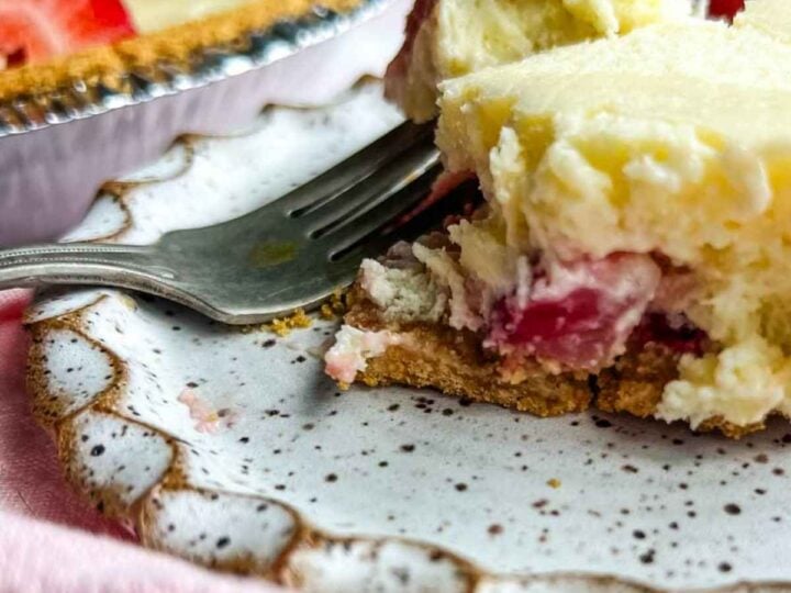 A close-up of a creamy strawberry cheesecake dessert on a plate with a fork and a pie in the background.