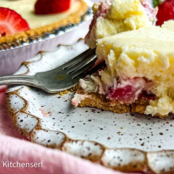A close-up of a creamy strawberry cheesecake dessert on a plate with a fork and a pie in the background.
