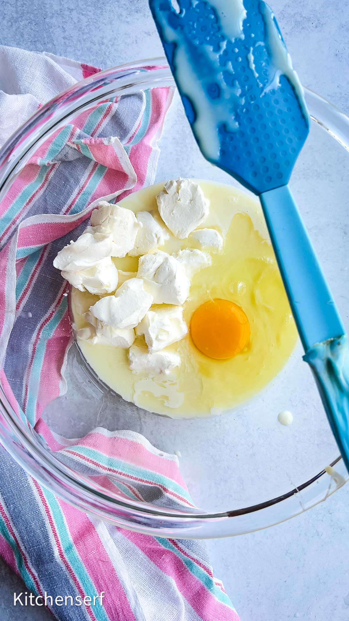 Egg, cream cheese, and sweetened condensed milk in a glass bowl with a blue spatula and striped towel on the side—perfect for starting a strawberry cheesecake.
