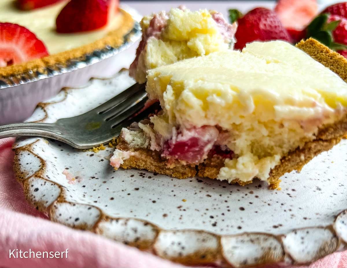 A fork cuts into a creamy strawberry cheesecake pie slice on a decorative plate, with strawberries in the background.