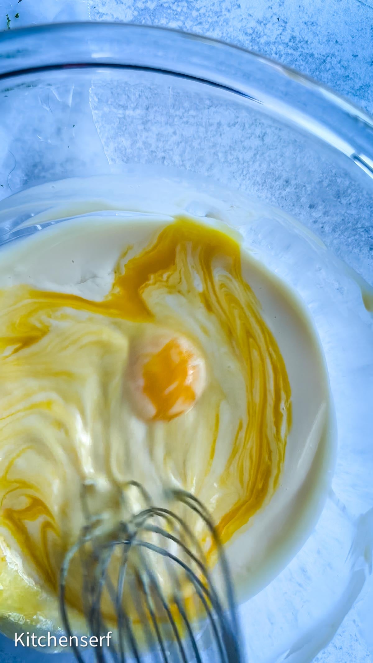 A clear bowl with eggs and batter, perfect for Nellie & Joe's Key Lime Pie, being mixed with a metal whisk on a light blue surface.