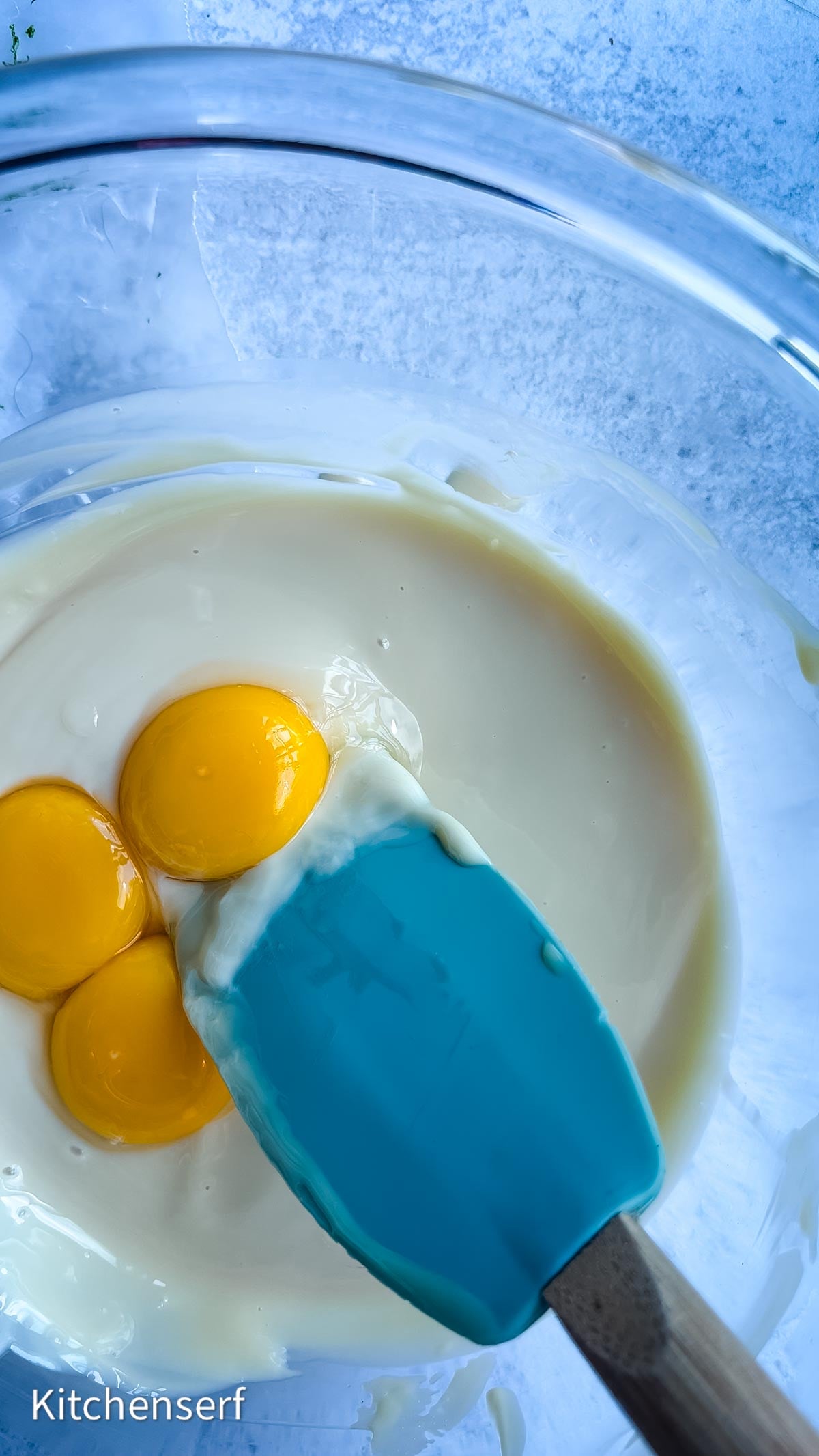 Three egg yolks in a bowl of batter with a blue spatula resting beside them, ready to be transformed into a classic Nellie & Joe's Key Lime Pie.