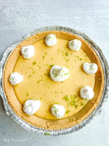 Key lime pie in a foil pan, made with Nellie & Joe's Key Lime Pie recipe, topped with whipped cream dollops and lime zest, on a light surface.