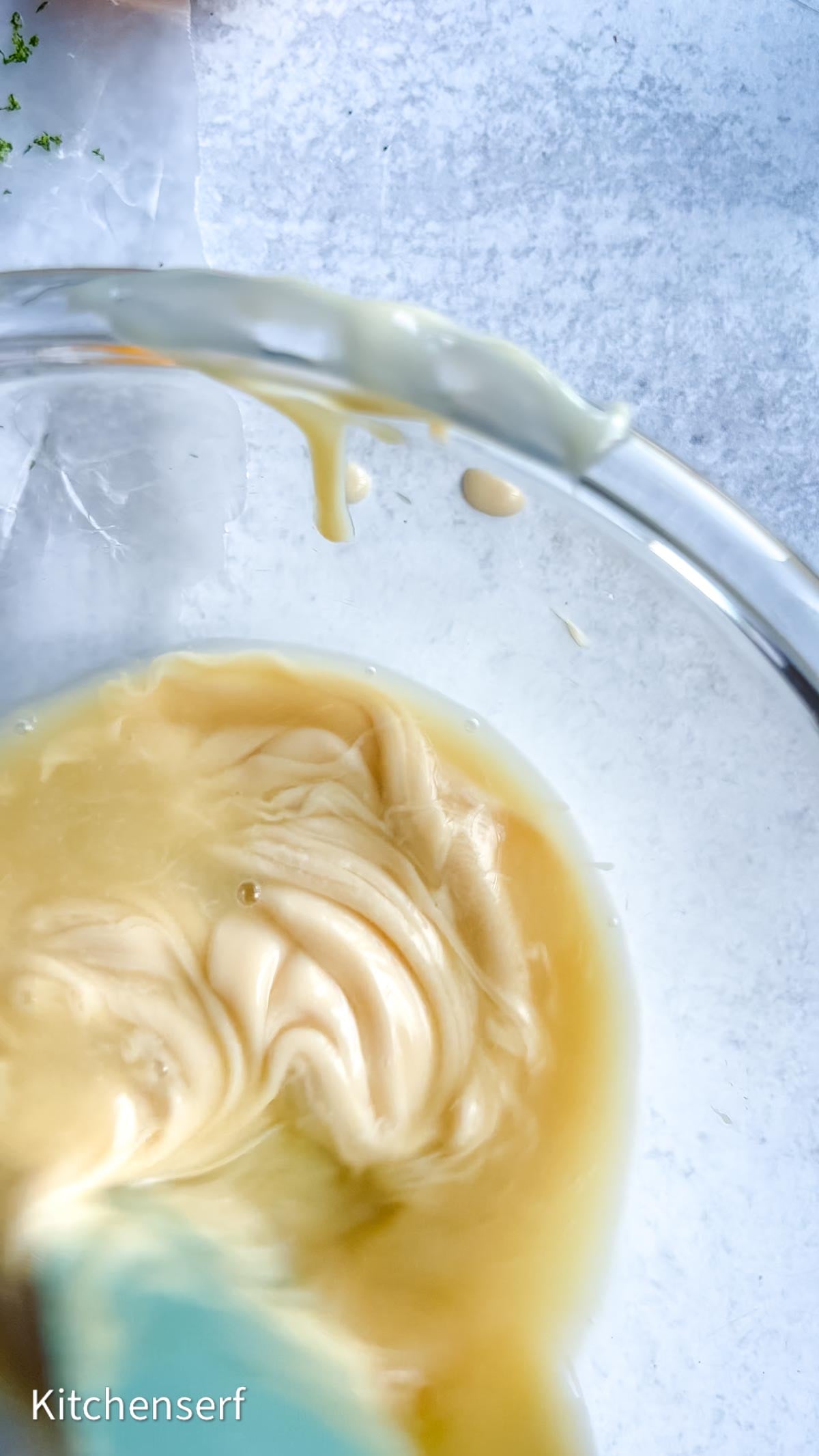 A close-up of a glass bowl with creamy Nellie & Joe's Key Lime Pie batter being stirred using a blue spatula.