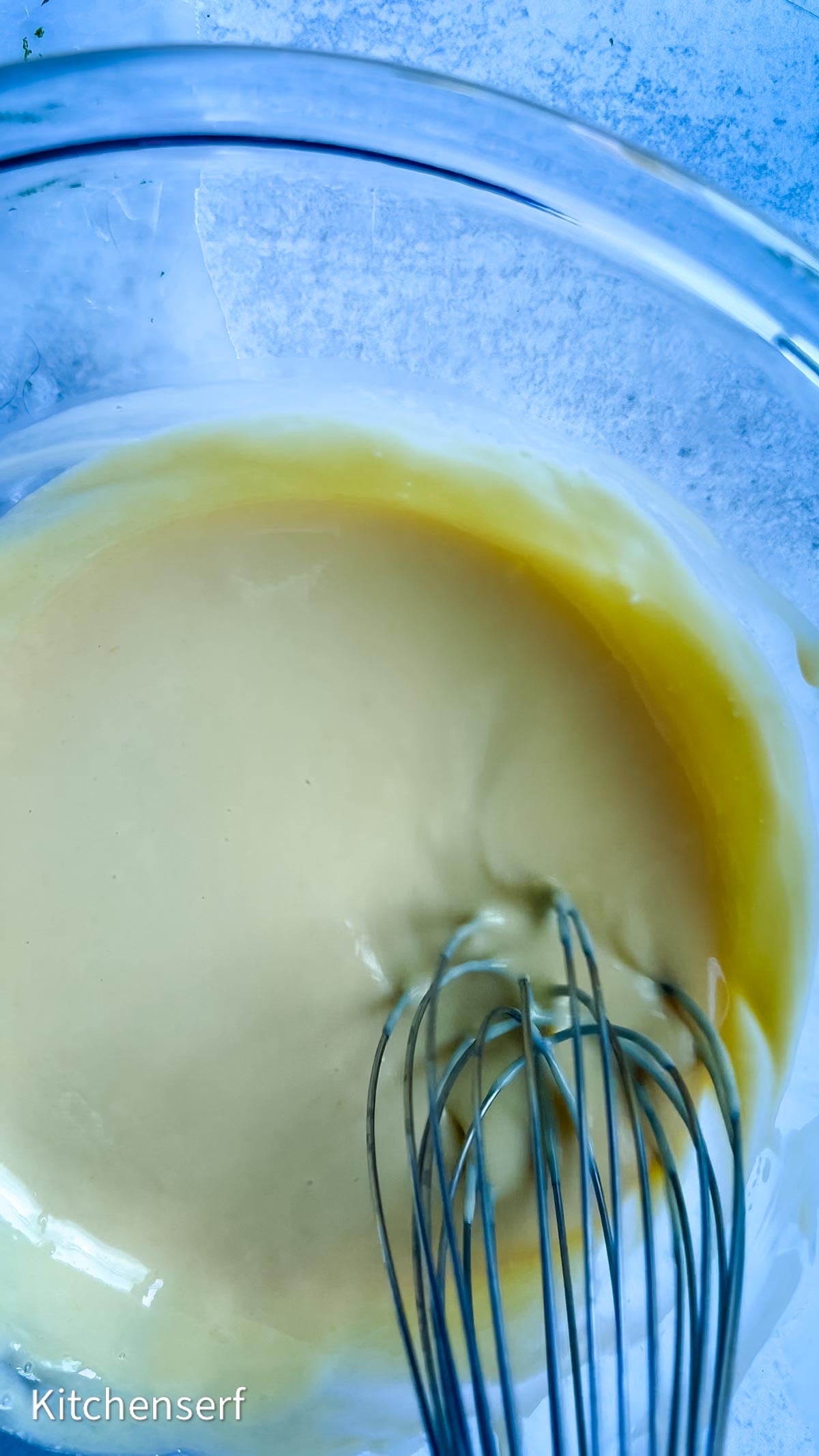 A metal whisk stirring creamy yellow Nellie & Joe's Key Lime Pie batter in a clear glass bowl on a blue surface.