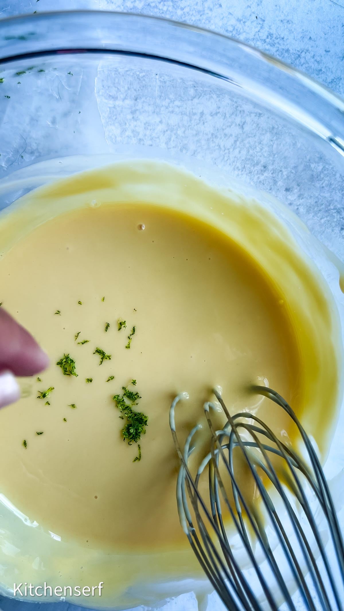 A hand whisks yellow batter in a glass bowl, with lime zest sprinkled on top, preparing the base for a classic Nellie & Joe's Key Lime Pie.
