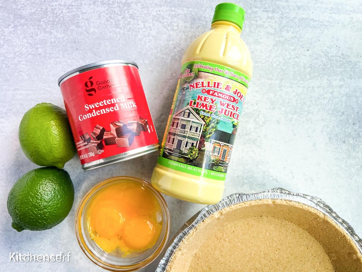 A pie crust, two limes, egg yolks, condensed milk, and a bottle of Nellie & Joe's Key Lime Pie juice are arranged on a gray surface.