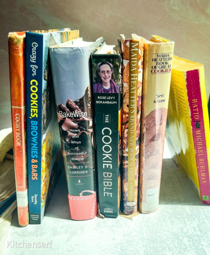 Seven baking books and cookie cookbooks stand upright on a sunlit kitchen counter, their spines facing forward.