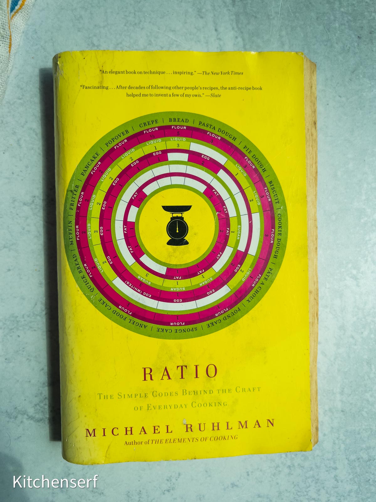 A yellow-covered book titled Ratio by Michael Ruhlman, one of the essential baking books, lying on a light blue surface.