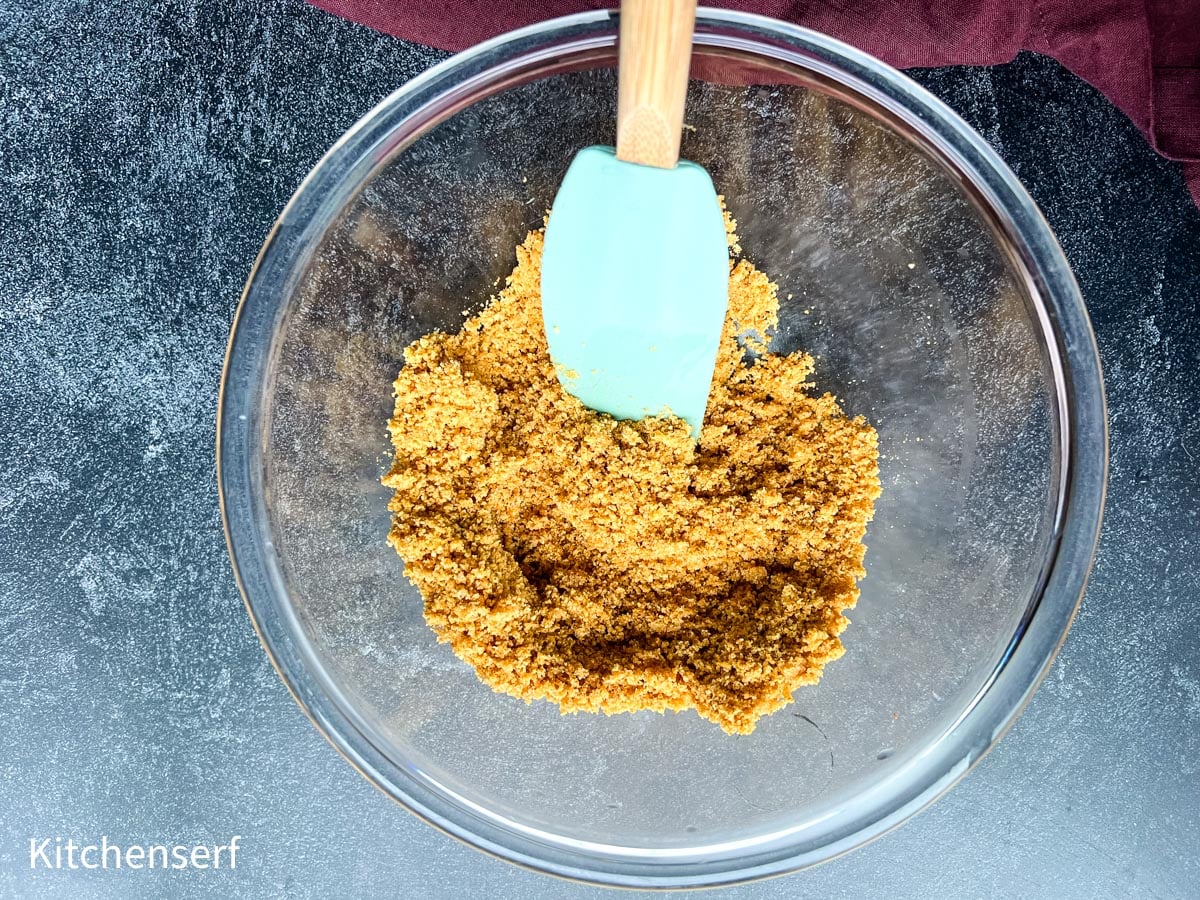 A spatula mixes crushed graham crackers in a glass bowl on a dark countertop, preparing the base for delicious smores bars.