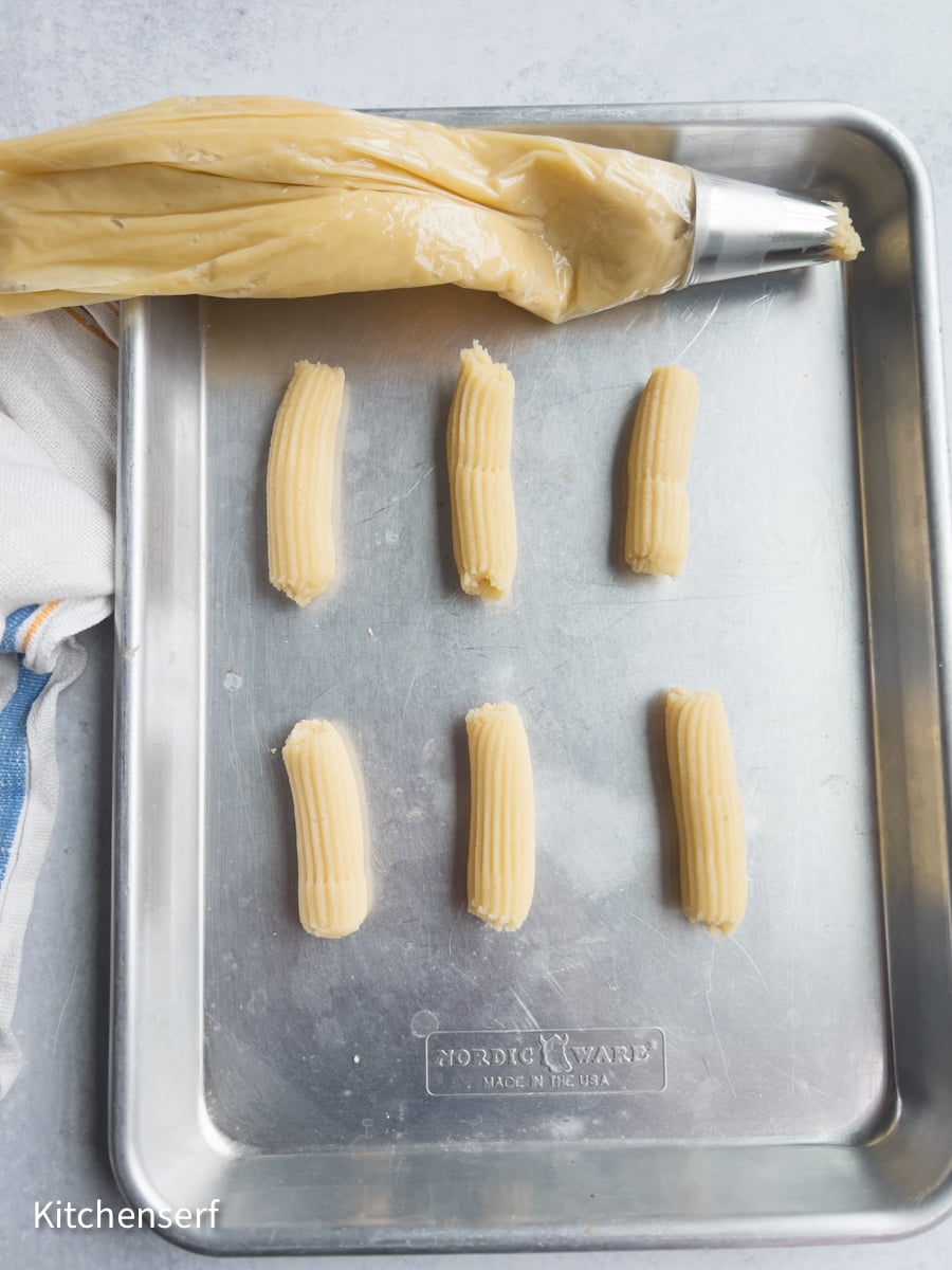 Piping bag and six piped dough strips for Italian butter cookies on a metal baking sheet, ready to bake.