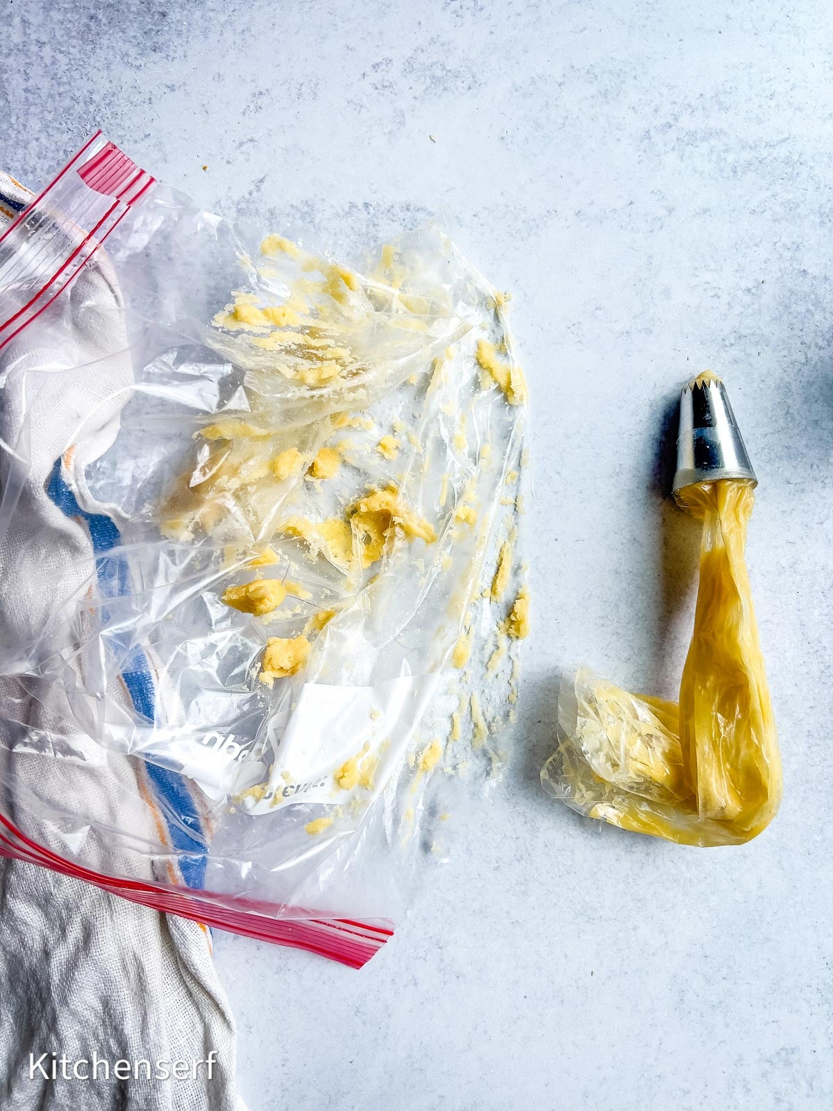 A messy plastic bag with dough, a cloth, and a piping bag fitted with a metal tip on a light gray surface.