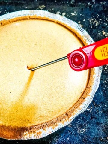 A thermometer reads 183.6°F as it checks the center of a pumpkin pie with in a foil crust on a dark surface.