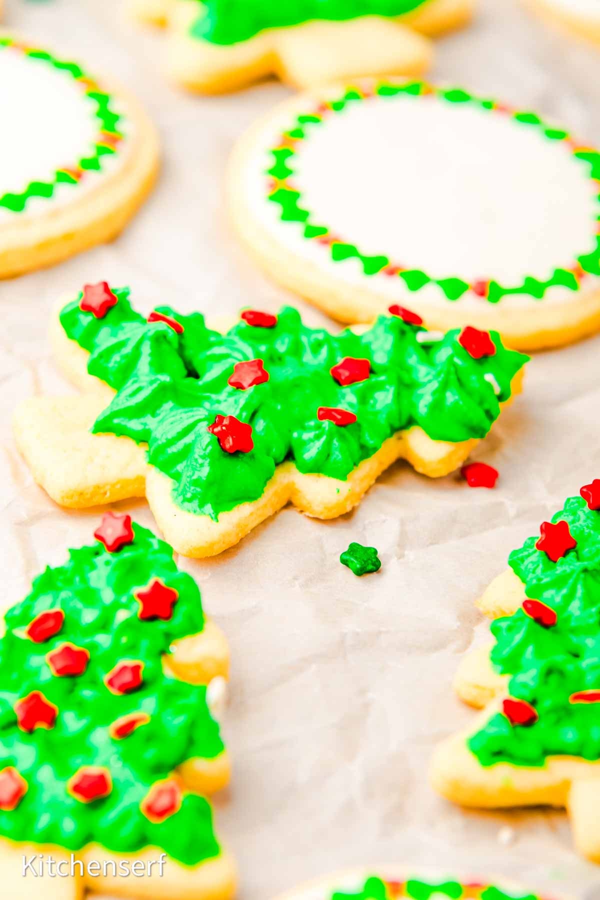 No spread cut-out cookies shaped like Christmas trees, decorated with green icing and red star sprinkles, arranged on parchment paper.