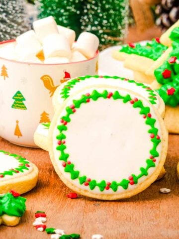 No spread cut-out cookies decorated with green and red icing, with a festive mug of marshmallows and Christmas trees in the background.