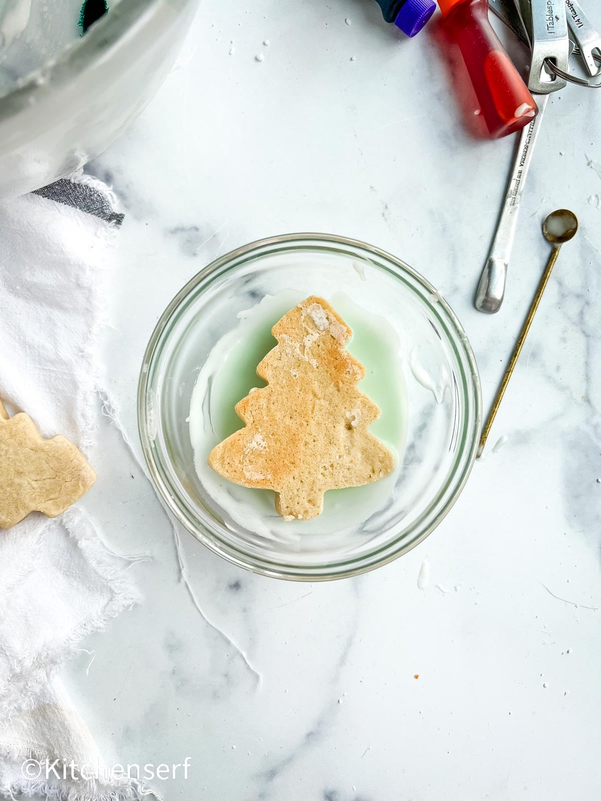 tree-shaped cookie in a bowl with icing remnants, surrounded by baking tools on a marble countertop.