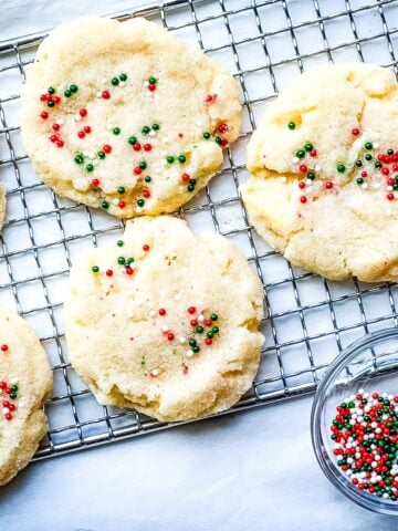 sugar cookies with sprinkles on top on a rack next to a small bowl of nonpareils.