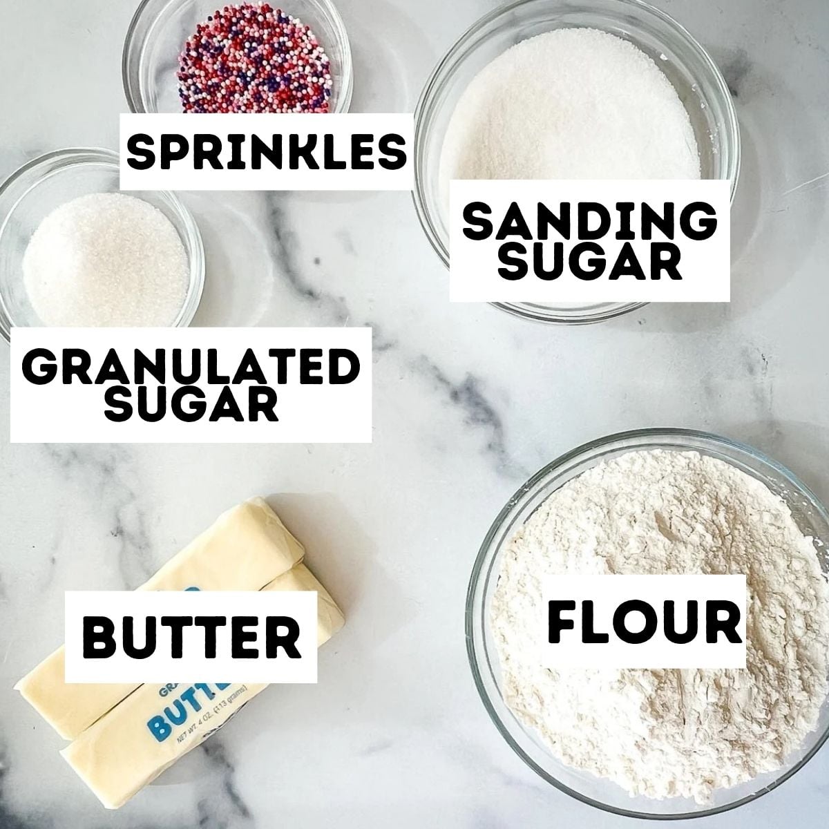 Bowl of sprinkles, sanding sugar, granulated sugar, flour, and sticks of butter on a white surface—perfect ingredients for making sugar cookies without baking powder.