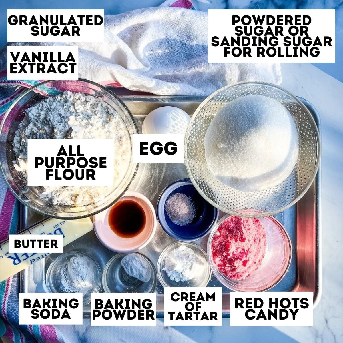A tray with labeled sugar cookie ingredients in bowls, including flour, eggs, butter, baking soda, and Red Hots for making festive red hots cookies.