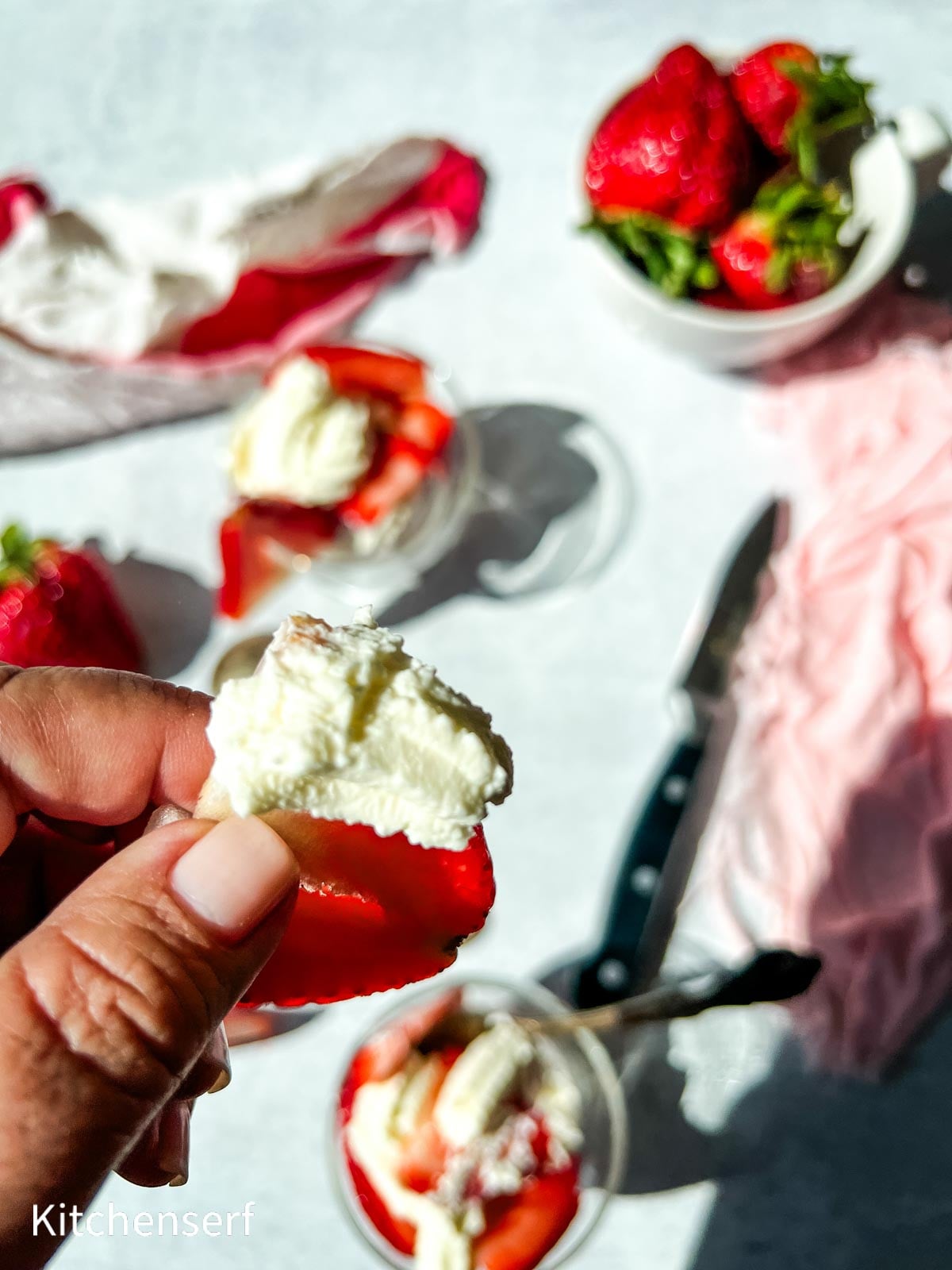 A hand holds a strawberry topped with whipped cream; more strawberries and cream are in the background.