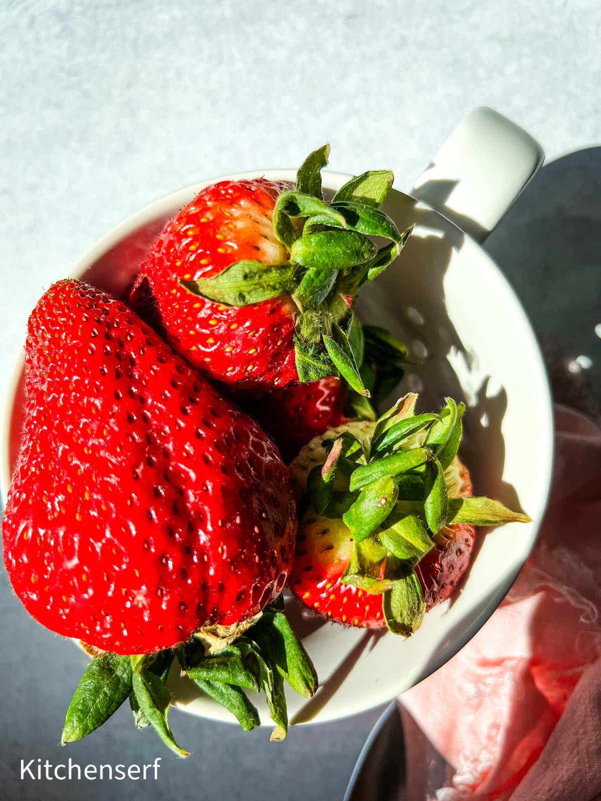 Three bright red strawberries with green stems in a white mug, with strong natural light and shadows.