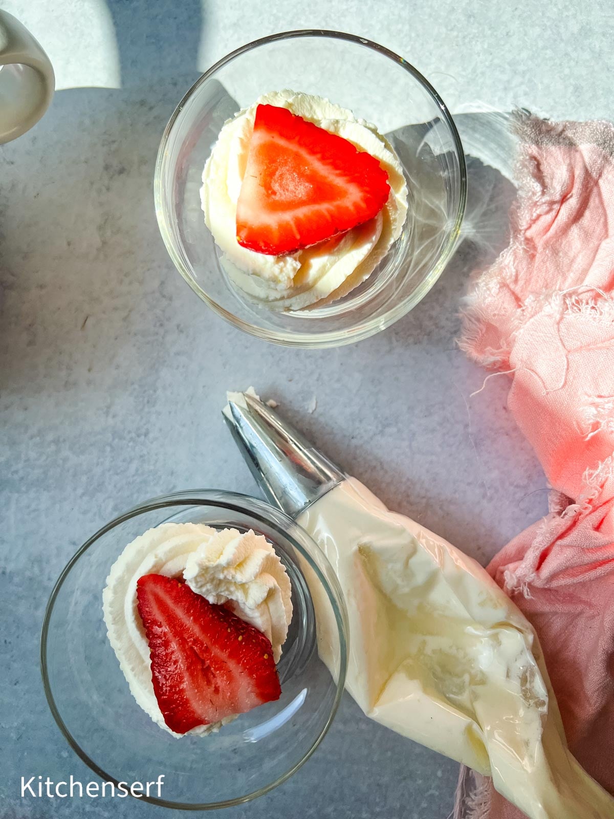 Two glass bowls with whipped cream and strawberry slices, pastry bag nearby, on a light surface with pink cloth.