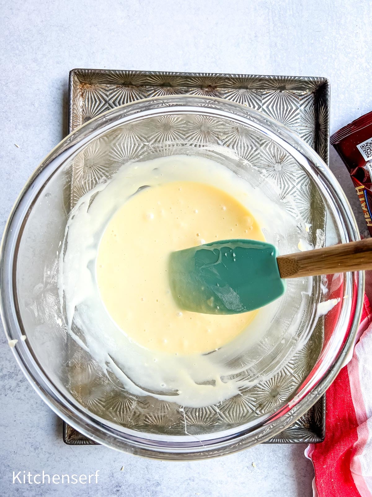 A glass bowl with creamy batter and a green spatula on a patterned metal tray.