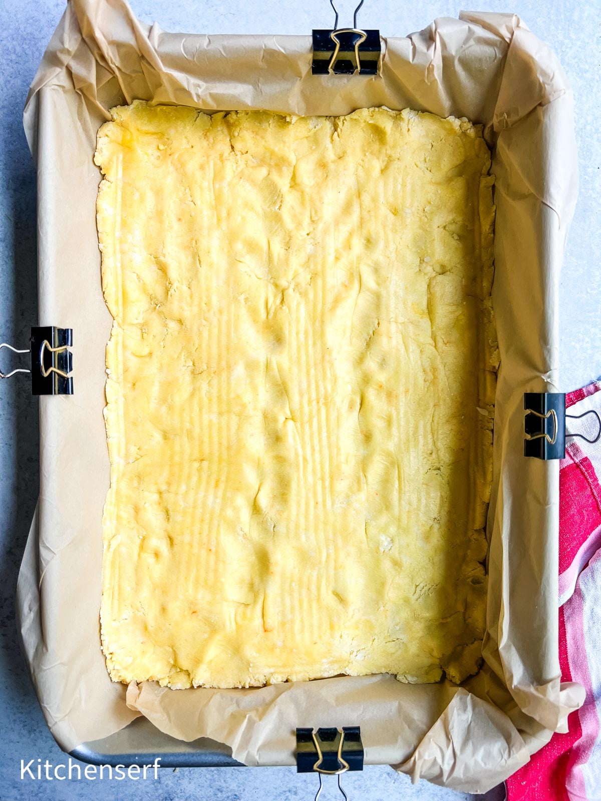 Baking pan lined with parchment and binder clips, filled with pressed shortbread dough, ready to bake.