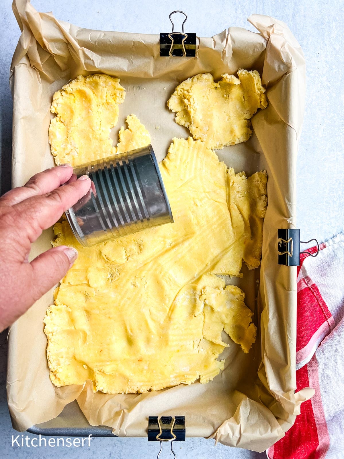 A hand uses a glass to press dough into a parchment-lined baking pan with binder clips.