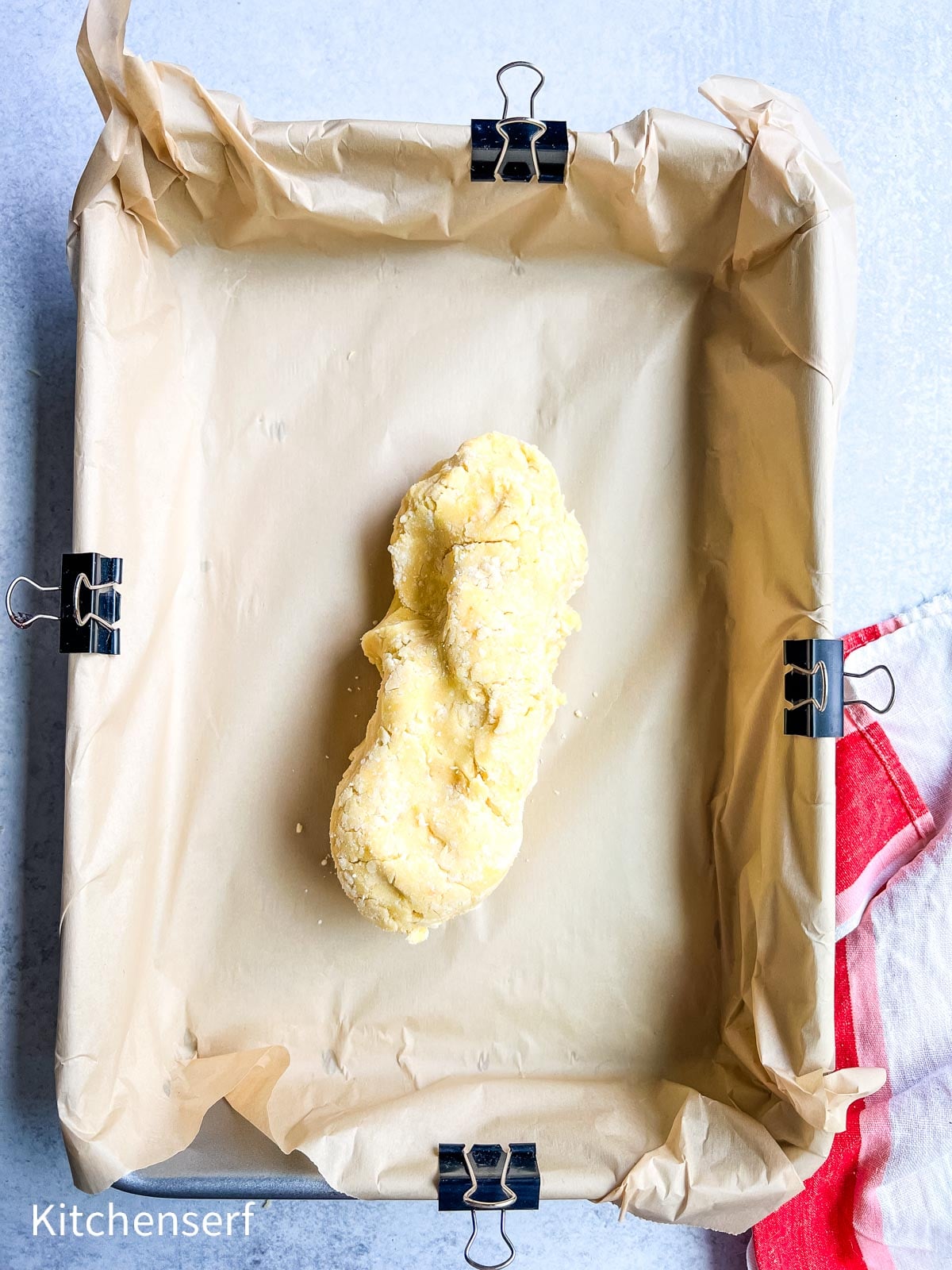 A lump of dough sits on parchment paper in a rectangular pan, secured with binder clips.