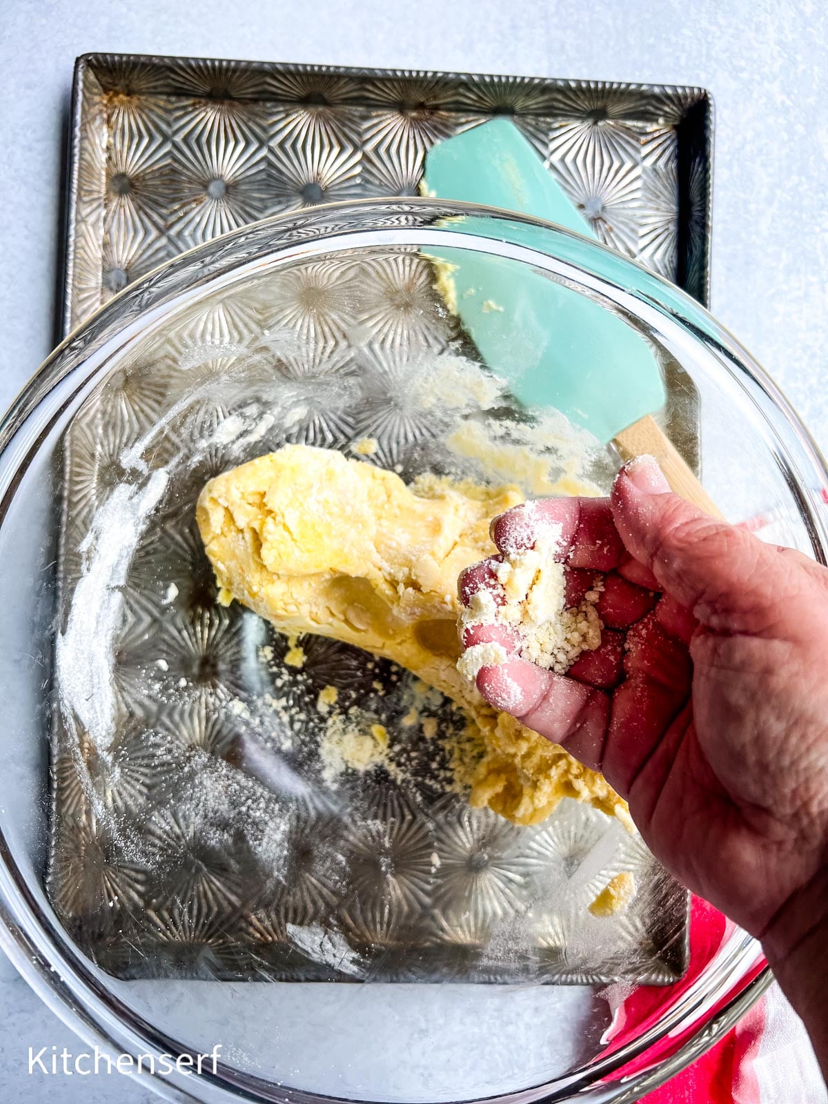 A hand sprinkles flour over dough in a glass bowl, with a baking tray and spatula nearby.