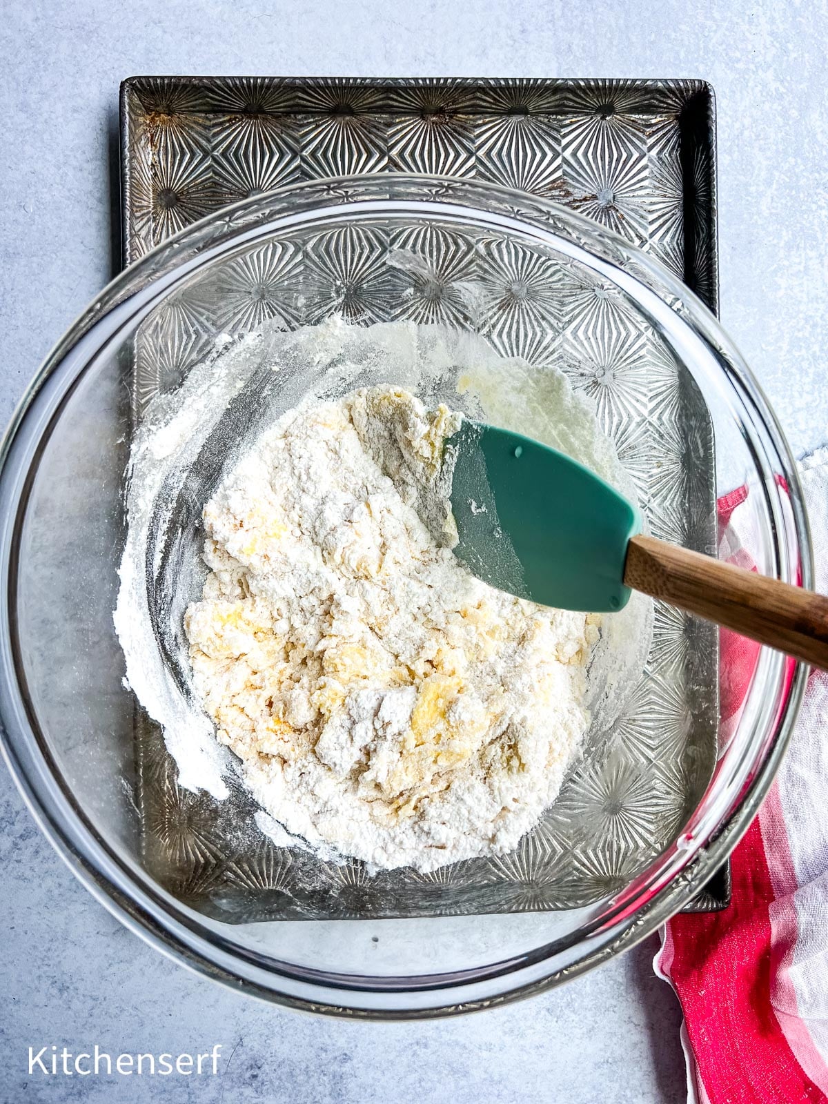 A glass bowl with flour mixture and spatula on a textured baking sheet, next to a red and white towel.